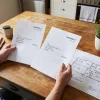 A Brighton homeowner compares painting proposals beside a floor plan and notebook while weighing recommendations and project details.