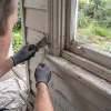 A painter examines peeling weatherboards beside a window, checking the timber condition before an exterior repaint.