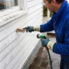 A painter washes coastal weatherboards beside a window as part of preparing the exterior for a new coating.
