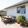 A white house wall, shaded window and grey roof edge sit beside patio furniture and potted plants.